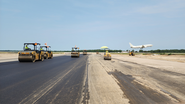 construction equipment paving runway with plane in background mid-takeoff