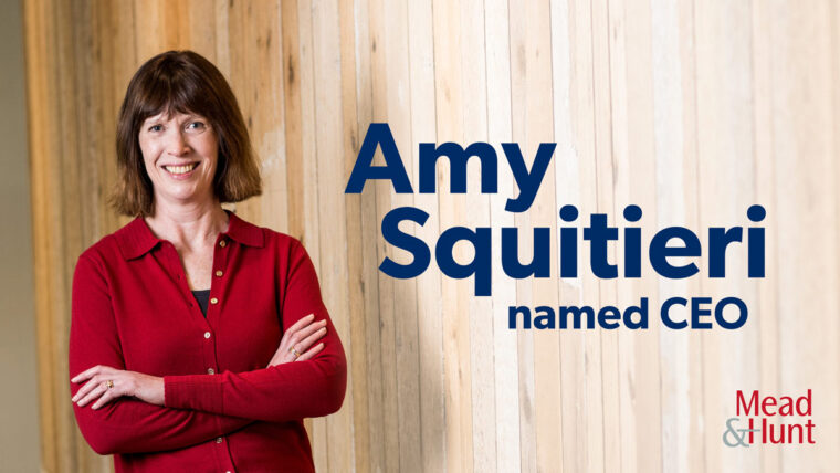 Amy stands in front of a wooden wall next to the words Amy Squitieri named CEO