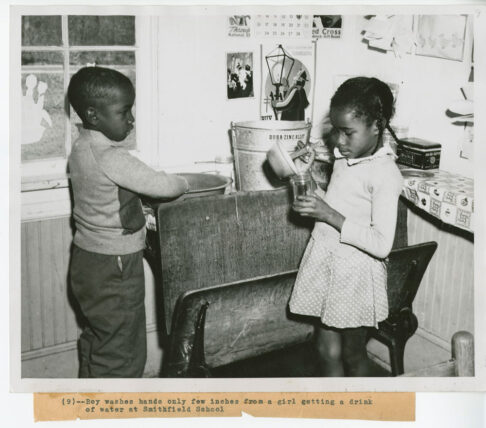Black and white photo of a young boy washing his hands in close proximity to a girl pouring water into her jar.