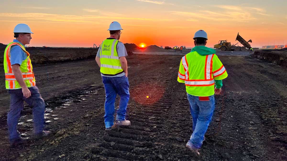 three people in high visibility gear and hard hats walk toward a sunset on an airfield
