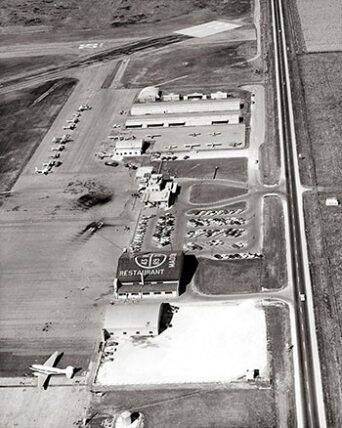 black and white aerial view shows a small airport, runway, aircraft, and building marked restaurant