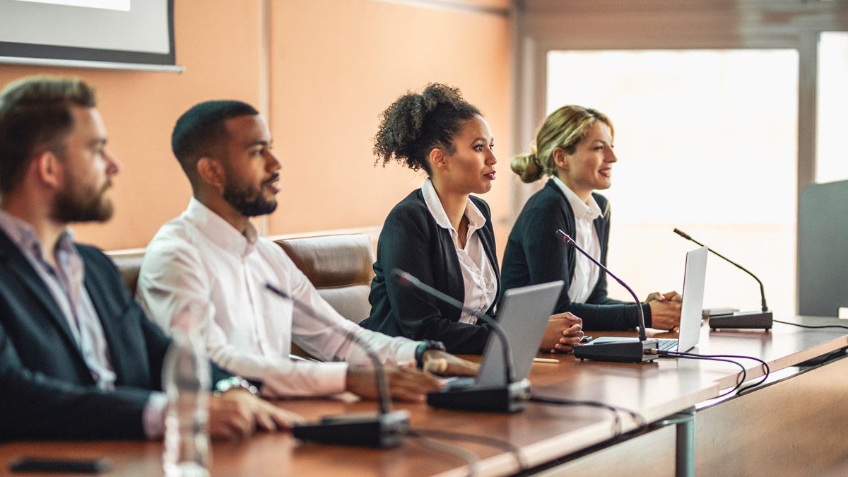 four executives sit behind a board table with laptops ready to speak