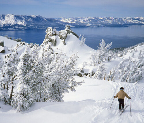 Person skiing down white snowy mountain overlooking Alpine Lake Tahoe and mountains in the distance