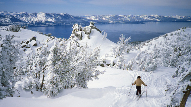 Person skiing down white snowy mountain overlooking Alpine Lake Tahoe and mountains in the distance