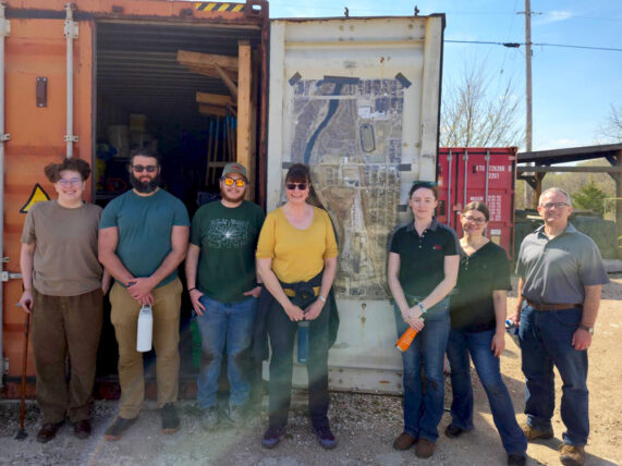 Amy stands centered by her team outdoors near an open shipping container