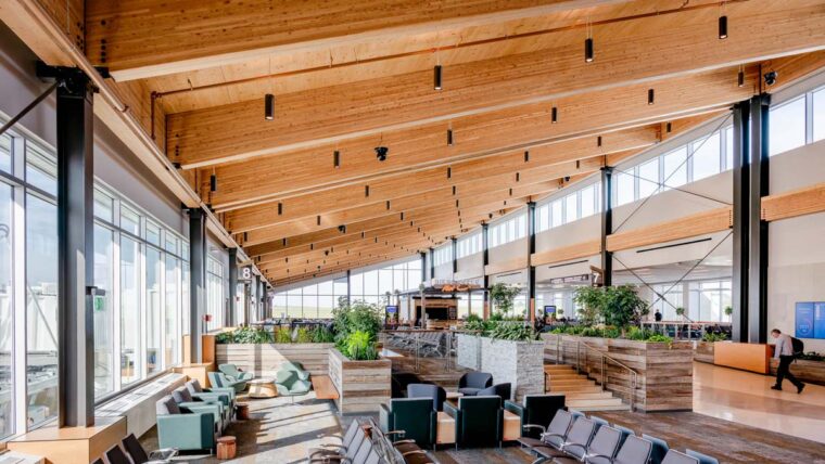 • Sunlit airport terminal with exposed mass timber beams spanning the roof above passenger seating and landscaped gathering spaces.