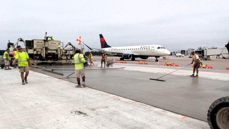 a team finishes concrete on the runway with an airplane taxiing nearby