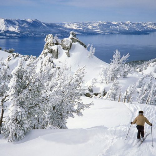 Person skiing down white snowy mountain overlooking Alpine Lake Tahoe and mountains in the distance