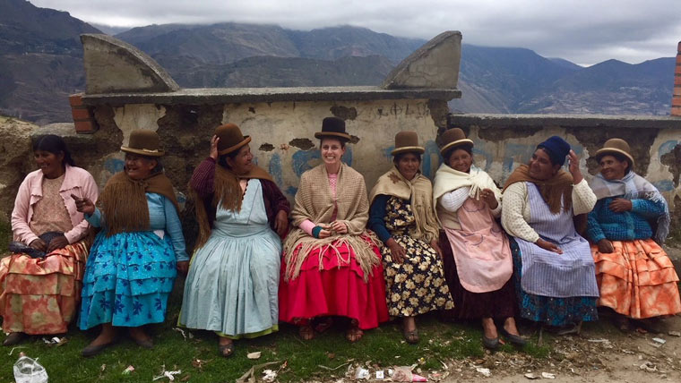 eight women sit in front a wall with mountains in the background. they are wearing traditional peruvian dresses in colorful prints. with brown shawls and hats.