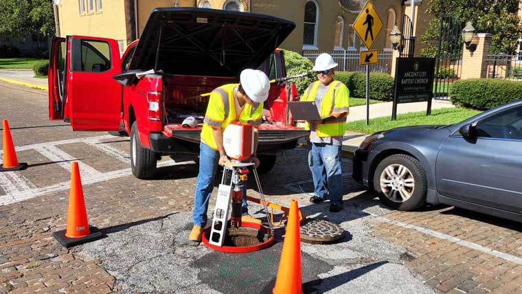 two workers wear hardhats and high-vis gear while working on a manhole in a road