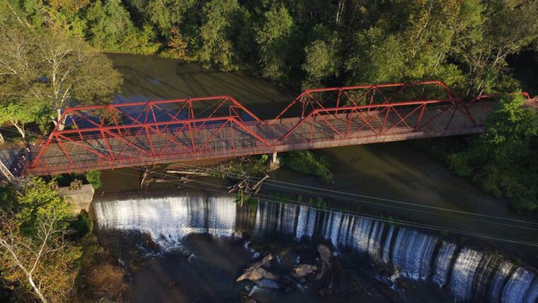 an aerial view of a pedestrian bridge over small manmade falls. The Bridge is red metal and wood.