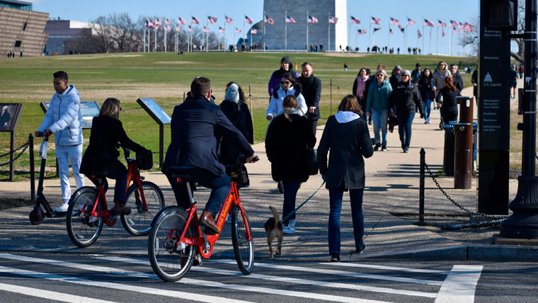 two bicyclists, a ma on a scooter, a woman with a dog and other pedestrians use a crosswalk in front of the Washington Monument.