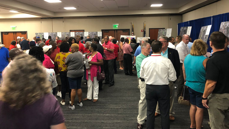a crowd stands in a large room at a public meeting. Posters line the perimeter.
