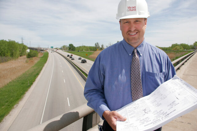 a younger John Rathke stands with a draft document on a bridge.