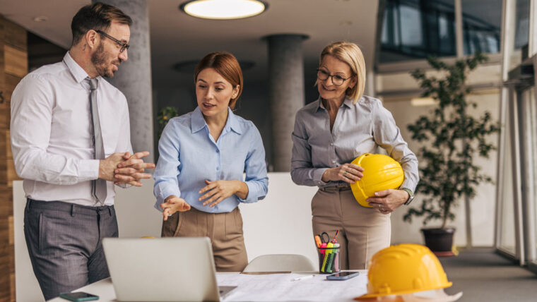 Smiling engineers and resilience planners gather around a worktable set with blueprints and a hard hat in a building where warm sunlight slants through tall windows.
