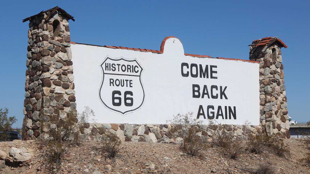 a large sign has the Route 66 sign with come back again in big black letters