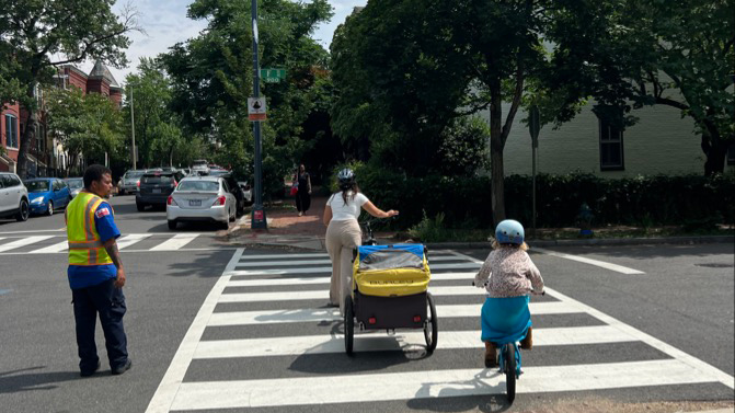 a crossing guard looks on as a woman and child on bikes cross an urban street