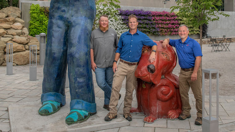 The project team poses next to the sculptures in Slack Plaza, including a brown dog and the legs of a sculpture so tall you can't see its upper half